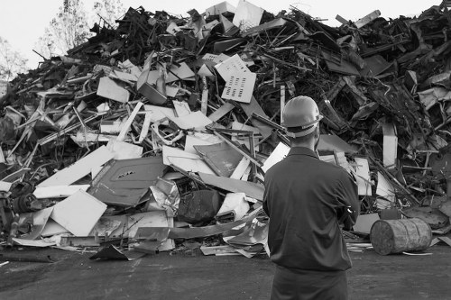 A man wearing a helmet and dark work clothing stands facing away from the camera, observing a large pile of mixed waste materials. The waste pile contains broken and discarded household appliances, plastic containers, metal fragments, and wooden debris, all piled haphazardly on a paved surface. The background shows a clear sky with some trees visible in the distance, suggesting an outdoor location near a waste disposal or recycling area. The environment appears industrial or municipal, with the focus on waste management activities. The scene emphasizes the scale of rubbish that a professional rubbish removal service such as Commercial Waste Harlesden manages, highlighting the variety and volume of waste materials typically handled in urban or suburban settings, including areas near Harlesden in London.