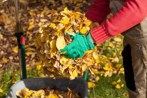 A person wearing a red long-sleeved garment and green gloves is shown collecting a large bundle of yellow and brown fallen autumn leaves. The leaves are being gathered from a garden or park area and are being held above a black plastic container or waste bin, which is positioned on a grass surface. In the background, there are additional leaves scattered on the ground, along with some gardening tools, including a leaf rake with a red handle. The scene suggests a leaf clearance activity typical of garden waste management in the Harlesden area, with the individual preparing to dispose of the collected foliage as part of rubbish removal or garden maintenance services provided by Commercial Waste Harlesden.