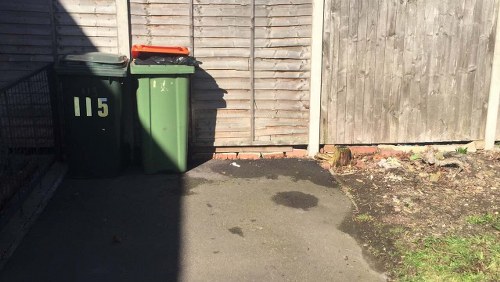 The image depicts two large, wheeled rubbish bins positioned on a paved surface in front of a wooden fence. The bin on the left is dark green with the number '115' painted in yellow on its front and has a closed black lid. The bin on the right is a lighter shade of green with a closed lid. The pavement shows a dark patch, possibly a stain, near the base of the bins. In the background, the wooden fence features vertical panels with a section of horizontally arranged panels on the right, and the overall environment suggests a residential or commercial exterior yard, typical for rubbish collection points in Harlesden. To the left, part of a metal gate or fence is faintly visible, enclosing the area. The scene is illuminated by daylight, casting shadows from the bins onto the ground, emphasizing the outdoor setting for waste management services in the area.