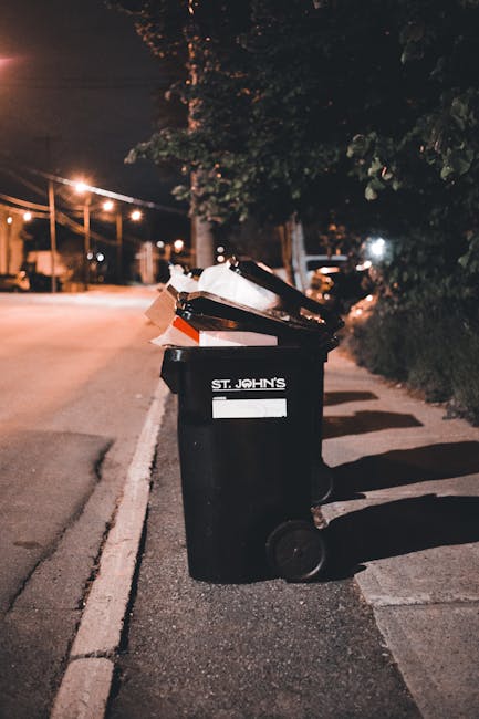 A black wheeled rubbish bin labeled 'St. John's' is positioned on a sidewalk at night, filled with various discarded items including cardboard boxes, plastic bags, and paper waste. The bin is situated
