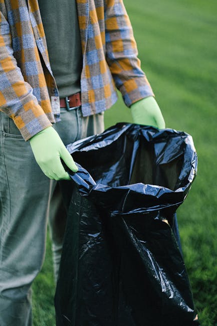 A black wheeled rubbish bin labeled 'St. John's' is positioned on a sidewalk at night, filled with various discarded items including cardboard boxes, plastic bags, and paper waste. The bin is situated