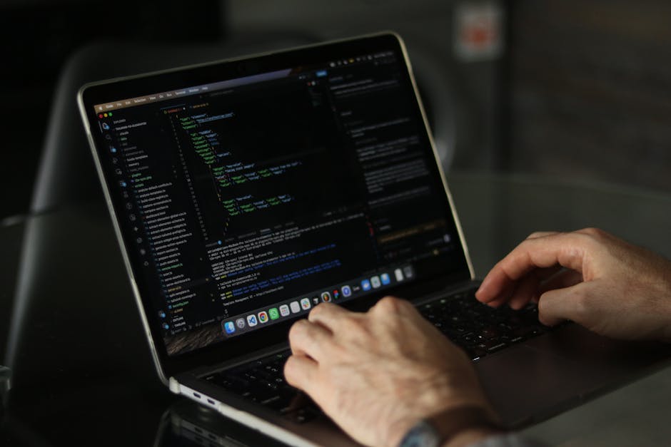 A person wearing a light blue shirt is seated at a desk, working on a laptop computer displaying lines of colorful programming code on the screen. The individual's left hand holds a pen, pointing towa
